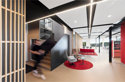 Offener Bürobereich im Verwaltungsbau Gläser in Horb mit Treppe, Lounge-Sitzmöbeln und roten Farbakzenten. Architektur arc+